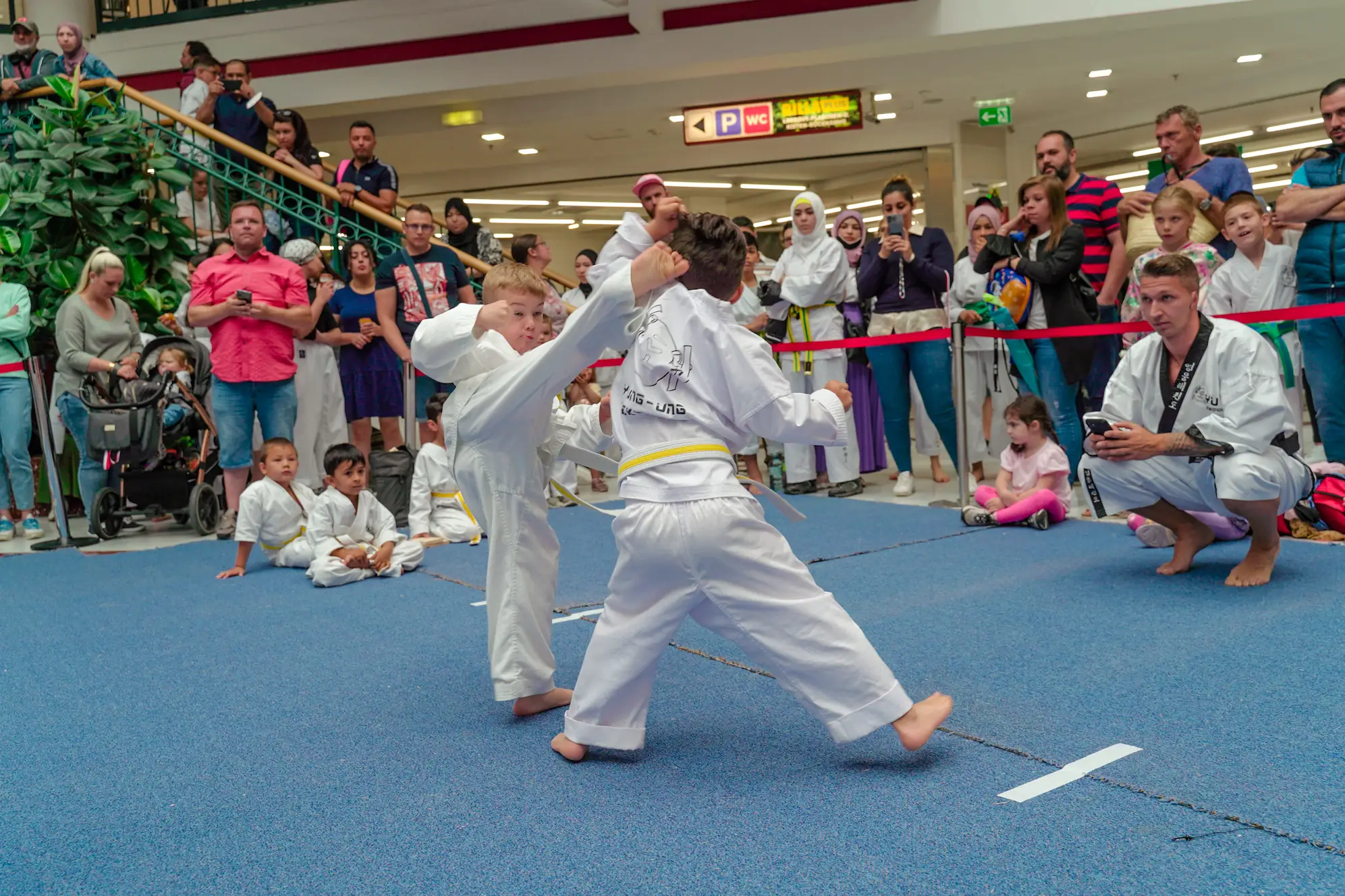 Zwei Kinder beim Taekwondo-Sparring auf einer blauen Matte vor Zuschauern in einem Einkaufszentrum.