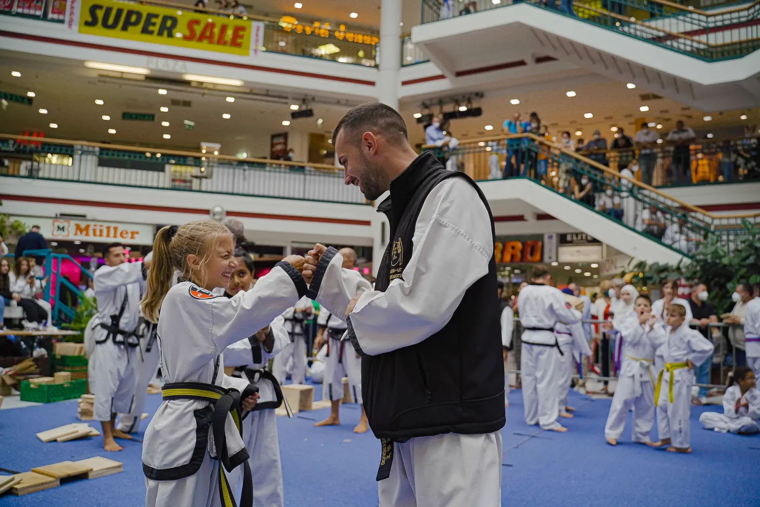 Lächelndes Mädchen und Trainer beim Faustgruß während einer Taekwondo-Vorführung in einem Einkaufszentrum.