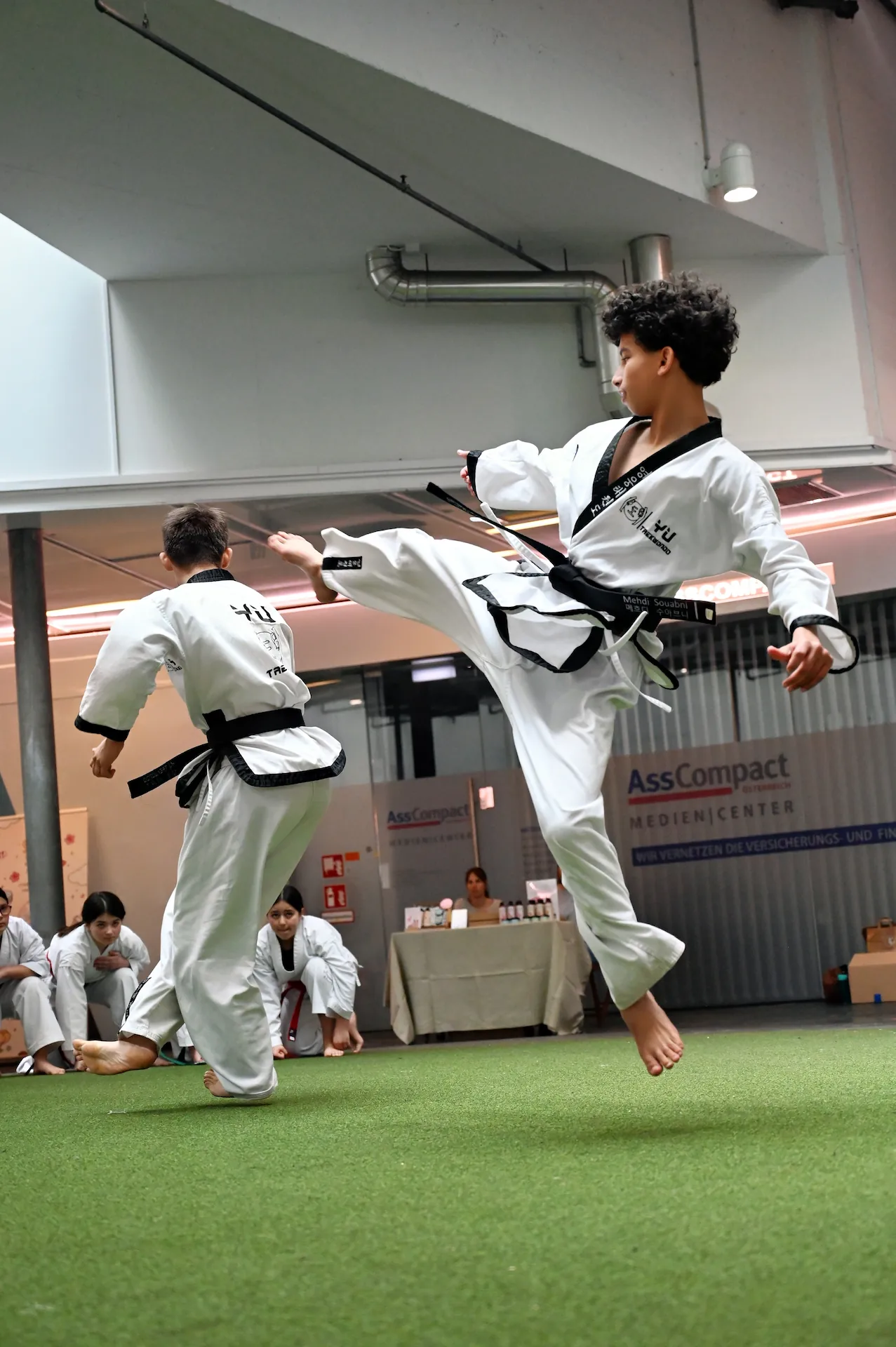 Ein Taekwondo-Kämpfer in der Luft führt bei einem Turnier einen spektakulären Tritt gegen seinen Gegner aus, während Zuschauer am Rand sitzen.